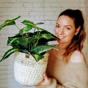 May include: A woman is holding a white crocheted plant pot with a green leafy plant inside. The pot has a brown leather tag attached to it. The woman is wearing a beige sweater.