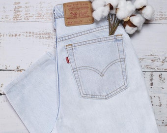 May include: Pale blue denim jeans with a classic five-pocket design, and a red tab on the back pocket. A brown leather patch is visible on the waistband. A bouquet of cotton stems rests near the jeans.