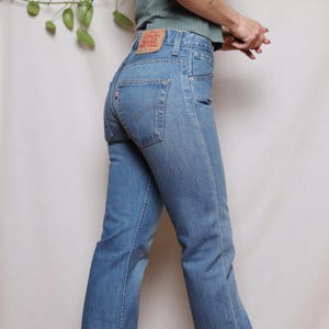 May include: Light blue denim jeans with a flared leg. The jeans have a classic five-pocket design, a red logo patch on the back, and a high waist. The jeans are a medium wash and have a vintage appearance.