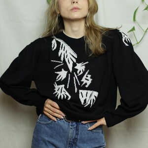 Può includere: Maglione nero con design floreale astratto bianco. Il maglione a maniche lunghe ha uno scollo tondo ed è abbinato a jeans blu. Il design è sulla parte anteriore e sulle spalle. La modella è davanti a uno sfondo bianco.