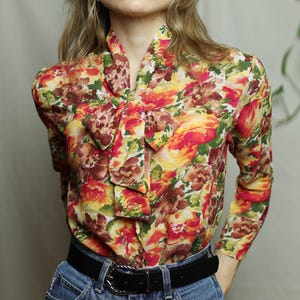 May include: A long-sleeved blouse with a floral print in shades of red, orange, yellow, and green. The blouse has a bow tie at the neck and a black belt.
