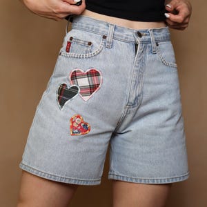May include: Light wash denim shorts with three heart-shaped patches. The patches feature plaid and floral patterns in red, black, and pink. The shorts have a classic five-pocket design and a button and zip closure.