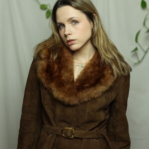 May include: A brown suede coat with a faux fur collar and a matching belt. The coat has a button-down front and a classic, vintage style. The collar is a lighter shade of brown than the coat itself.