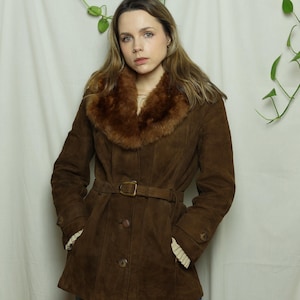 May include: A brown suede coat with a faux fur collar and a matching belt. The coat has a button-up front and long sleeves. The coat is a medium length and has a classic, vintage style.