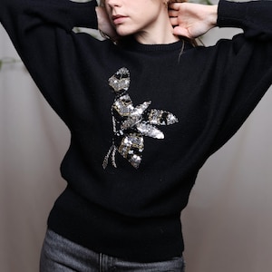 Può includere: Maglione nero con un motivo floreale. Il maglione ha un girocollo e maniche lunghe. Il motivo floreale è composto da paillettes e perline argentate e dorate. Il maglione è realizzato in morbido tessuto a maglia.