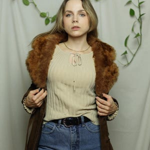 May include: A woman wearing a brown suede coat with a faux fur collar. The coat is open, revealing a beige ribbed knit top and blue jeans. A black belt with a silver buckle is visible.