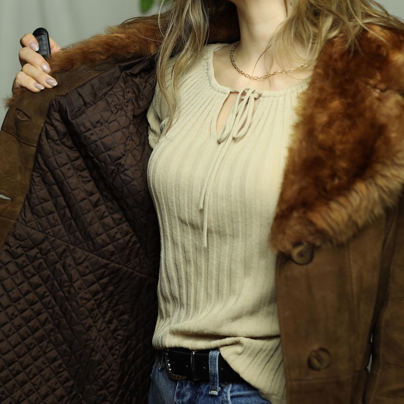 May include: Brown suede coat with a faux fur collar and quilted lining. The coat has large buttons and a classic design. The interior lining is a dark brown color. The coat is open, revealing the interior.