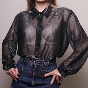 May include: A long-sleeved, sheer black button-down shirt featuring a subtle, abstract white scratch-like pattern throughout the fabric. The shirt has a classic collar, a front button placket, and buttoned cuffs, worn tucked into dark blue denim jeans.