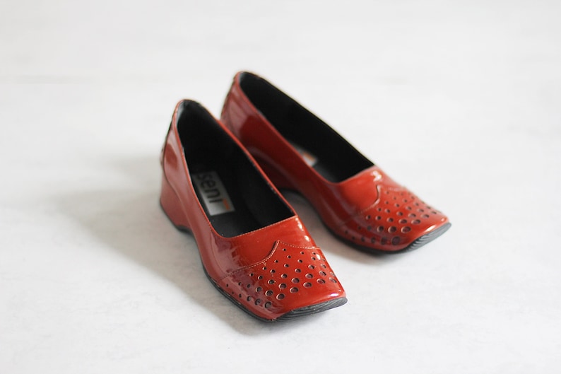 May include: A pair of red leather shoes with a small wedge heel and a perforated design. The shoes have a black lining and the brand name "Seni" is visible on the inside of one shoe.