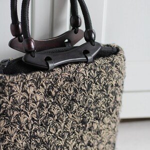 Puede incluir: Un peque&ntilde;o bolso de mano negro y beige con una textura tejida &uacute;nica. El bolso tiene dos asas negras con detalles de madera y una cremallera negra.