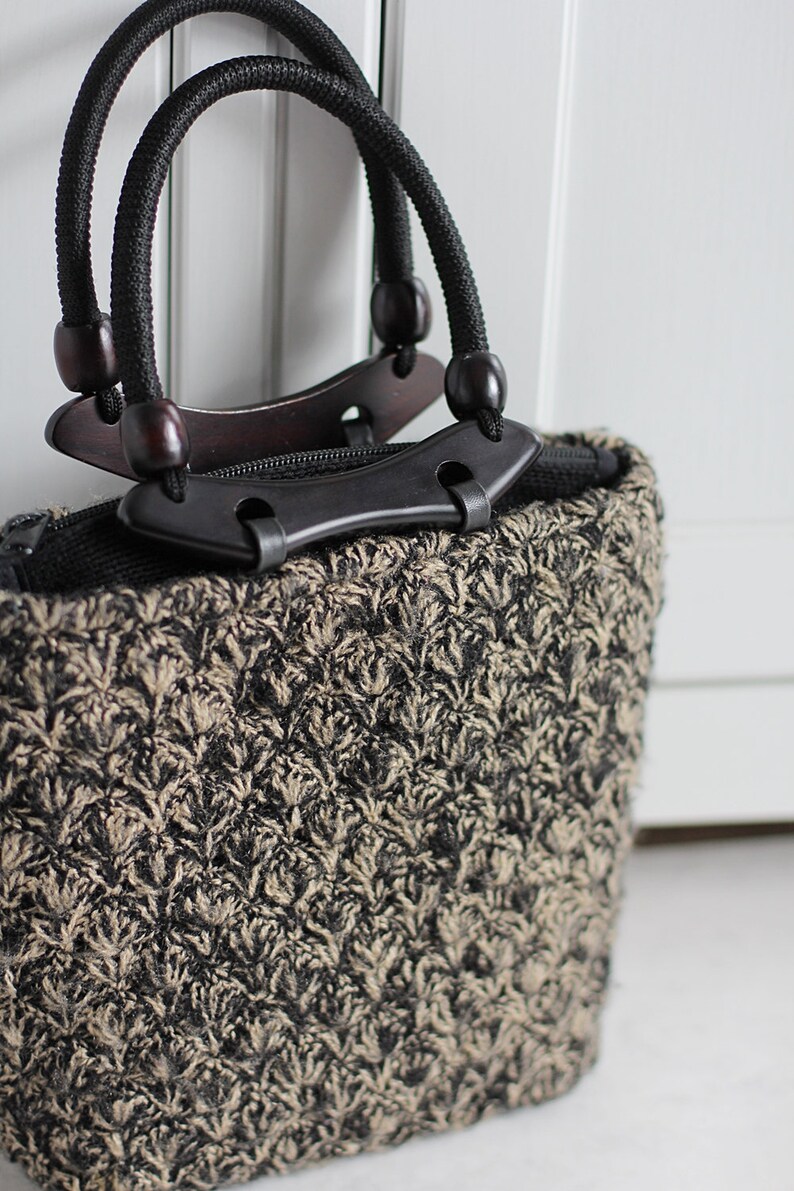 Puede incluir: Un peque&ntilde;o bolso de mano negro y beige con una textura tejida &uacute;nica. El bolso tiene dos asas negras con detalles de madera y una cremallera negra.