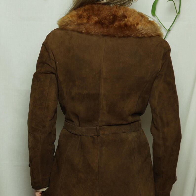 May include: A brown suede coat with a faux fur collar and a belt at the waist. The coat has long sleeves and a classic design. The fur collar is a lighter shade of brown than the coat.