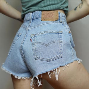 Puede incluir: Pantalones cortos de mezclilla azul claro con dobladillo deshilachado y un bolsillo trasero con una etiqueta roja que dice "Levi's".