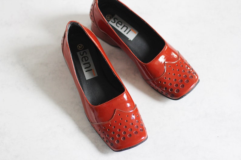 May include: A pair of red leather shoes with a perforated design and the word "seni" printed on the inside of each shoe.