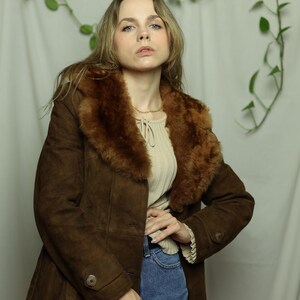May include: A brown suede coat with a faux fur collar. The coat has a button closure and a tailored fit. The model is wearing a cream-colored top and blue jeans. The coat is a vintage style.