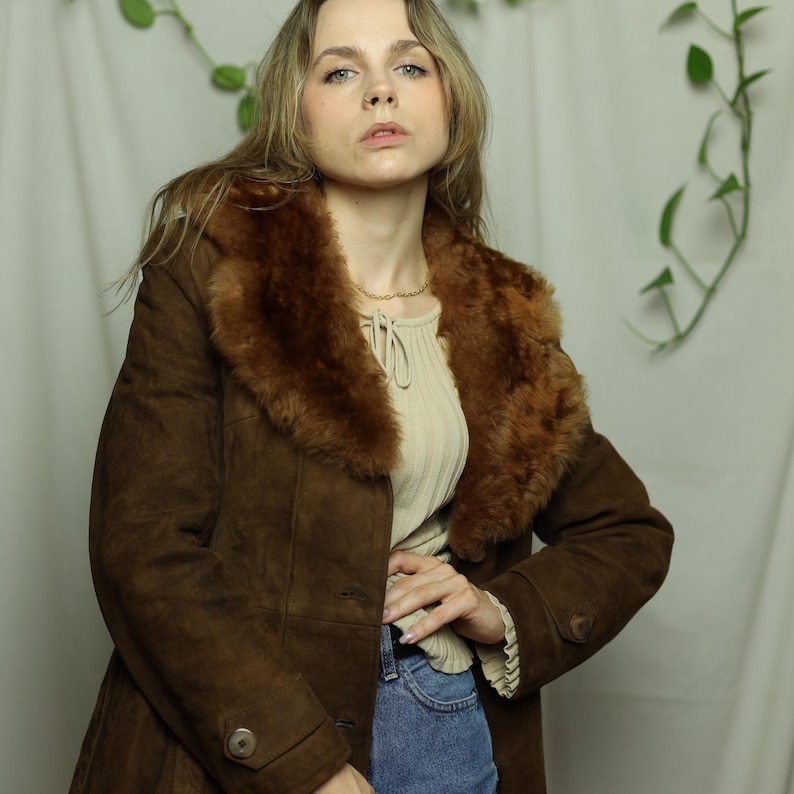 May include: A brown suede coat with a faux fur collar. The coat has a button closure and a tailored fit. The model is wearing a cream-colored top and blue jeans. The coat is a vintage style.