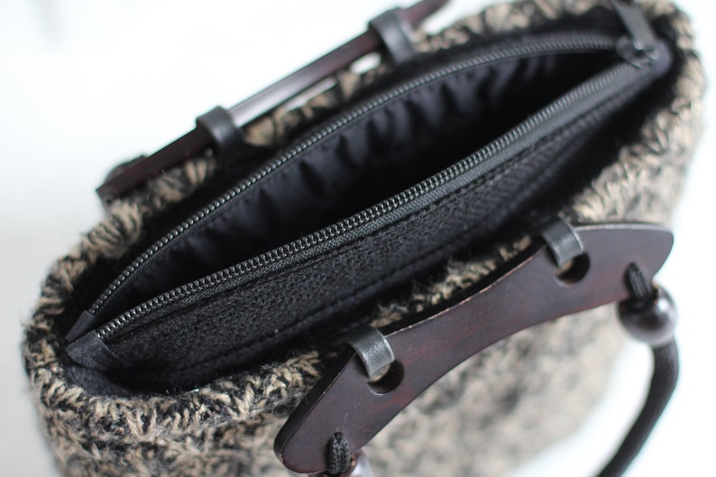 Puede incluir: Una bolsa negra con cremallera, con un asa de madera marr&oacute;n y un cord&oacute;n negro. La bolsa est&aacute; hecha de un tejido tejido con un patr&oacute;n blanco y negro.
