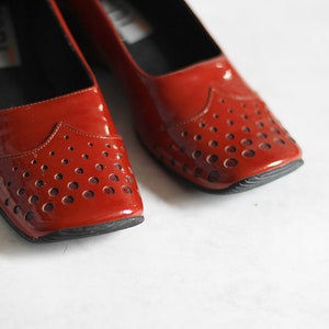 May include: A pair of red patent leather shoes with a perforated design. The shoes have a rounded toe and a low heel.