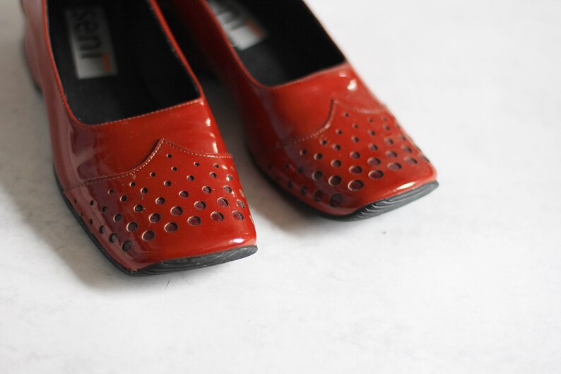 May include: A pair of red patent leather shoes with a perforated design. The shoes have a rounded toe and a low heel.