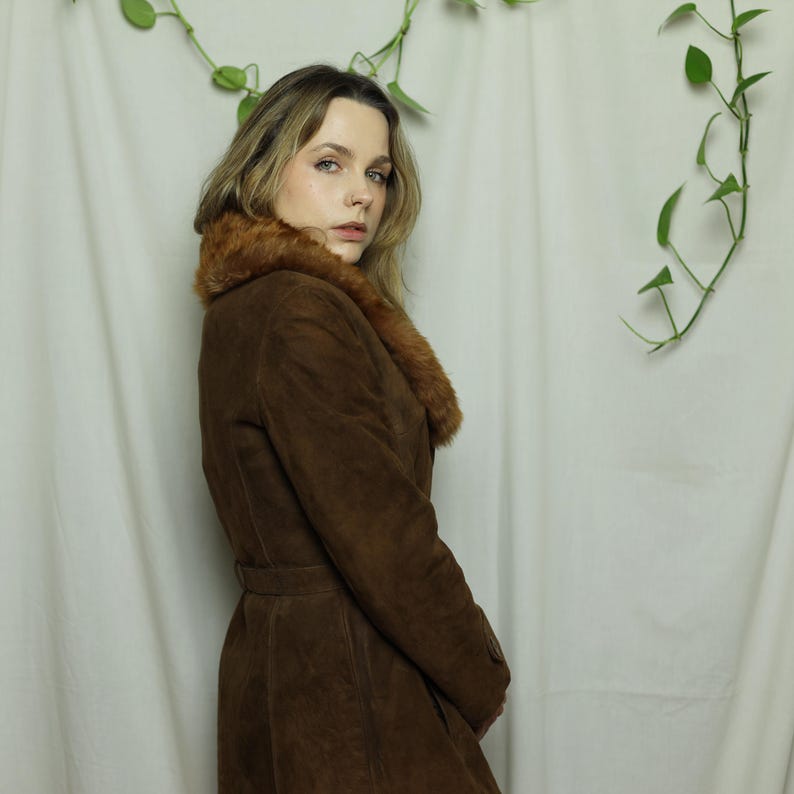 May include: A brown suede coat with a faux fur collar and a matching belt. The coat is knee-length and has a classic, vintage style. The fur collar is a lighter shade of brown, adding a touch of luxury to the coat.