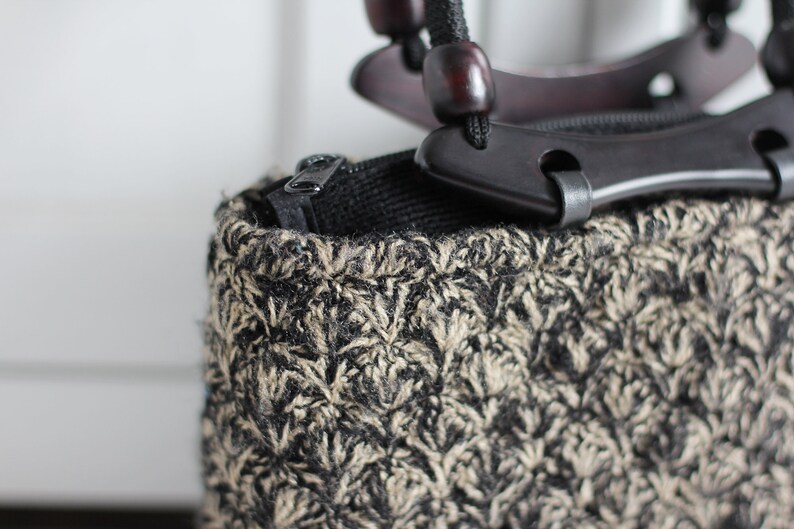 Puede incluir: Un bolso tejido negro y marr&oacute;n con una cremallera negra y un asa de madera con detalles negros.