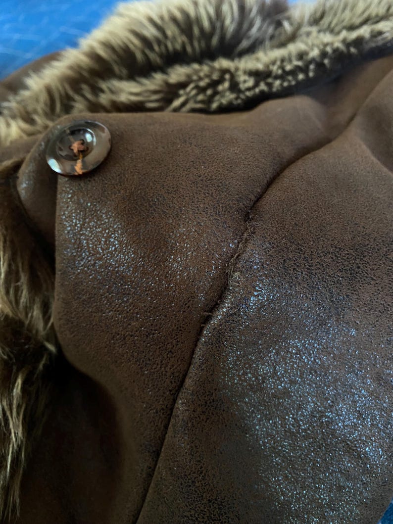 Pode incluir: Close-up de uma jaqueta de couro sint&eacute;tico marrom com gola forrada de pele. A jaqueta apresenta um bot&atilde;o grande e redondo e costuras vis&iacute;veis. A pele &eacute; uma mistura de cores marrom e bege.