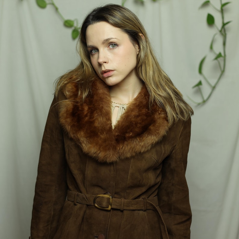 May include: A brown suede coat with a faux fur collar and a matching belt. The coat has a button-down front and a classic, vintage style. The collar is a lighter shade of brown than the coat itself.