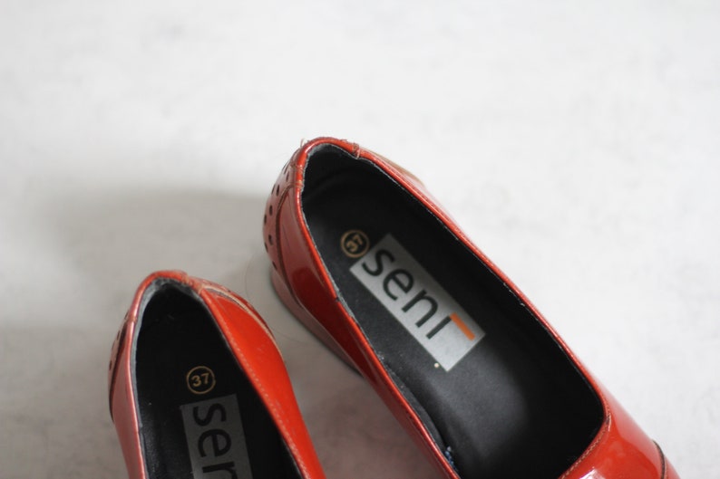 May include: A pair of red leather shoes with a black insole. The shoes have a small gold circle with the number 37 on the insole. The brand name "seni" is printed on the insole of the shoe.