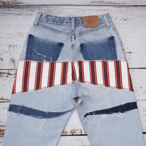 Op de afbeelding: Lichtblauwe spijkerbroek met een horizontaal paneel van witte, rode en donkerblauwe verticale strepen op de achterkant. De broek heeft donkerblauwe denim patches op de achterzakken en gerafelde, rauwe donkerblauwe denim accenten op de pijpen.
