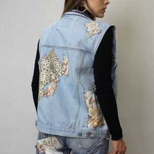 Può includere: Gilet in denim blu chiaro con toppe in stampa leopardata e tessuto floreale. Il gilet è abbinato a jeans coordinati, creando un ensemble in denim unico e rattoppato. Una maglia nera a maniche lunghe completa l'outfit.