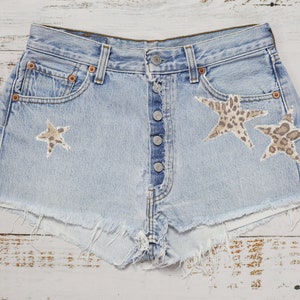 Op de afbeelding: Lichtblauwe spijkerbroekshorts met een knoopsluiting en een gerafelde zoom. De shorts hebben twee stervormige patches met een bruin en bruin luipaardprintpatroon. Een klassieke, casual stijl.