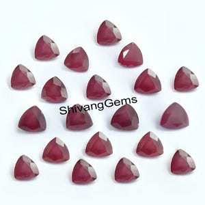 Può includere: Una collezione di pietre preziose di rubino triangolari e sfaccettate. Le gemme sono di colore rosso intenso e sono disposte su una superficie bianca. Il testo "Shivang Gems" è stampato in bianco.