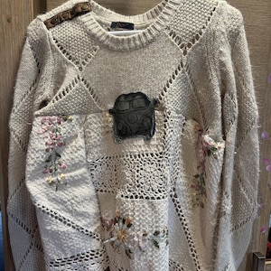 Peut inclure: Un pull beige à manches longues présentant un mélange de textures en tricot et en crochet. L'avant affiche un écusson gris foncé en forme de van, des broderies florales roses et vertes, et une patte d'épaule en similicuir marron à trois boutons.