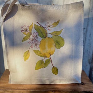 May include: A natural canvas tote bag featuring a watercolour illustration of a lemon branch. The design includes a yellow lemon, green leaves, and small pink flowers. The bag has two handles and is made of a natural-coloured fabric.