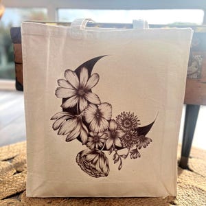 May include: A beige tote bag with a crescent moon design filled with detailed floral illustrations in brown tones. The bag has two white handles and is made of a sturdy fabric, suitable for carrying various items.