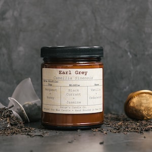 May include: A brown glass jar candle with a black lid and a white label that reads "Earl Grey Camellia Sinensis". The label also lists the scent notes: Bergamot & Honey, Black Currant & Jasmine, Vanilla & Cedarwood. The candle is sitting on a gray surface with loose tea leaves and tea bags.