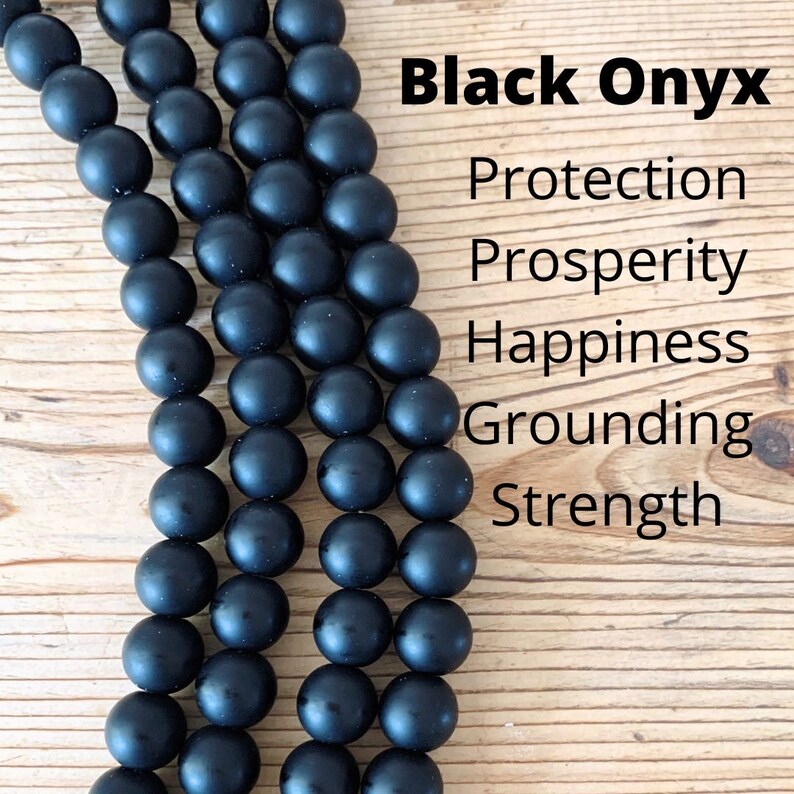 May include: Black Onyx beads strung together in a long strand. The beads are smooth and matte black. The text "Black Onyx" is written above the beads, and the text "Protection, Prosperity, Happiness, Grounding, Strength" is written below the beads.