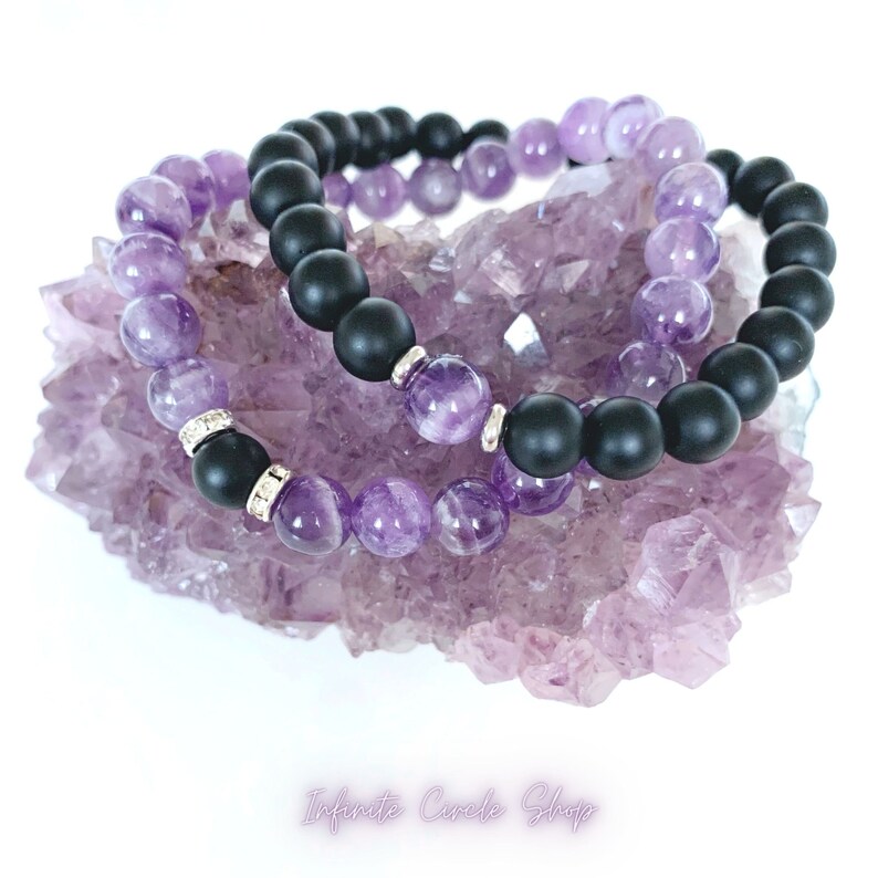 May include: Two bracelets, one with black matte beads and one with amethyst beads, are displayed on a purple amethyst cluster.