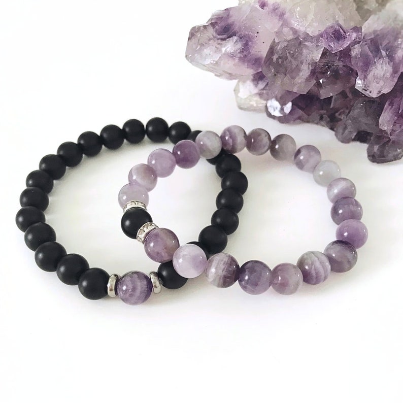 May include: Two bracelets, one black and one purple, made of beads. The purple bracelet has a gradient of light to dark purple beads.