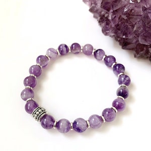 Pu&ograve; includere: Un braccialetto di ametista viola con perline d'argento e un ciondolo d'argento. Il braccialetto &egrave; su uno sfondo bianco.