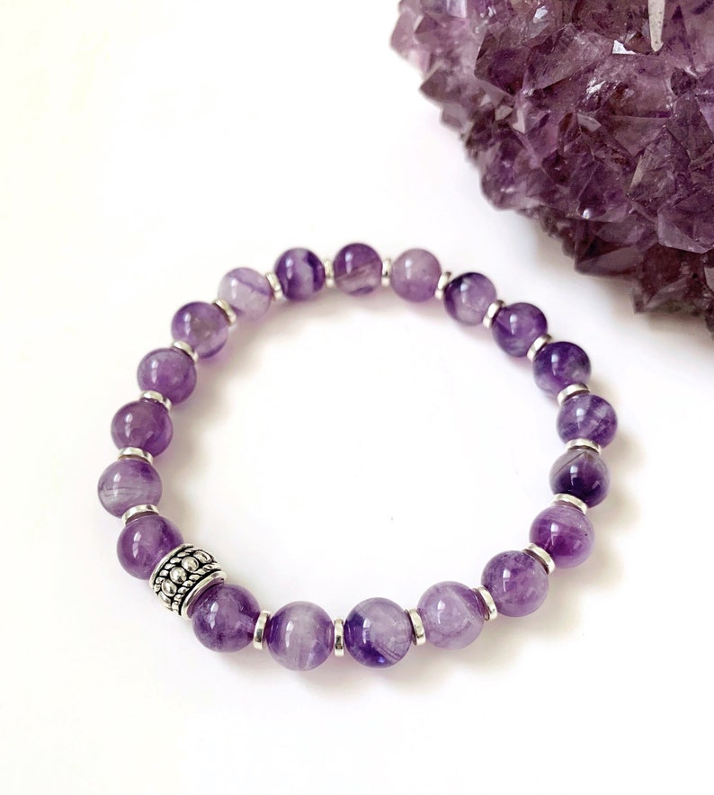 Pu&ograve; includere: Un braccialetto di ametista viola con perline d'argento e un ciondolo d'argento. Il braccialetto &egrave; su uno sfondo bianco.