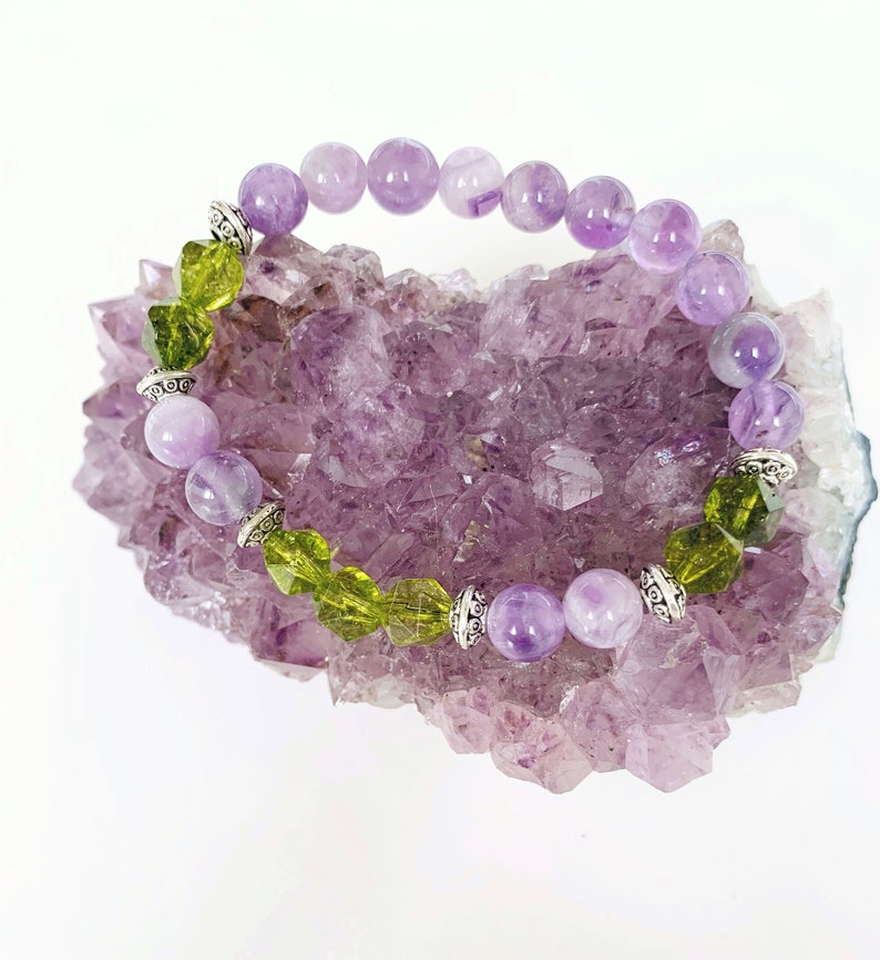 May include: A purple and green gemstone bracelet with silver accents. The bracelet is resting on a bed of purple amethyst crystals.