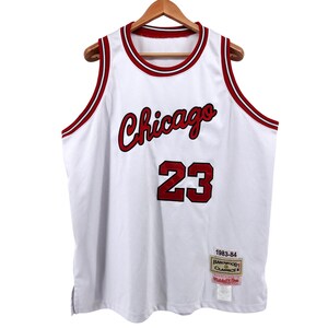 May include: White basketball jersey with red accents, featuring "Chicago" in script and the number "23". The jersey has red trim around the collar and armholes, and a "1983-84 Hardwood Classics" tag.