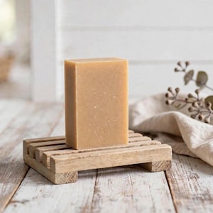 May include: A rectangular bar of light tan soap with visible flecks of texture, resting upright on a slatted wooden soap dish. The soap has a smooth, matte finish and a solid, uniform appearance, positioned centrally on the wooden surface.