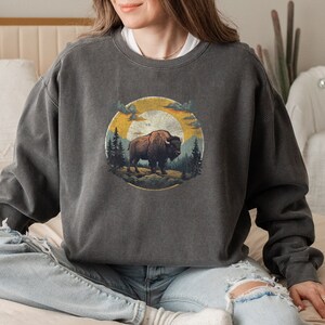A woman wearing a dark gray oversized sweatshirt featuring a detailed, vintage-style illustration of a bison standing on rugged terrain, surrounded by pine trees. The design is set within a circular frame, depicting a warm, golden sunset sky with clouds. She pairs the sweatshirt with light-wash, distressed jeans and cozy socks while sitting cross-legged on a neutral-toned bed. The soft, bohemian-inspired room setting enhances the relaxed and nature-inspired aesthetic of the outfit.