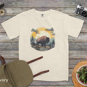 May include: A white t-shirt with a vintage-style illustration of a bison standing in a forest with a sunset in the background. The illustration is surrounded by a circle with a rustic woodgrain texture.