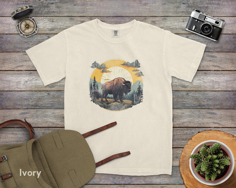 May include: A white t-shirt with a vintage-style illustration of a bison standing in a forest with a sunset in the background. The illustration is surrounded by a circle with a rustic woodgrain texture.