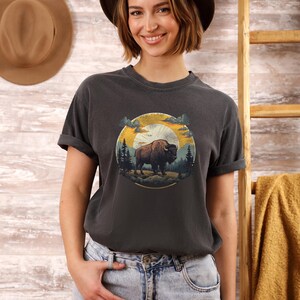 May include: A dark gray t-shirt with a vintage-style graphic of a buffalo standing in a forest with a sunset in the background. The graphic is in a circle with the words "Wild & Free" written below it.
