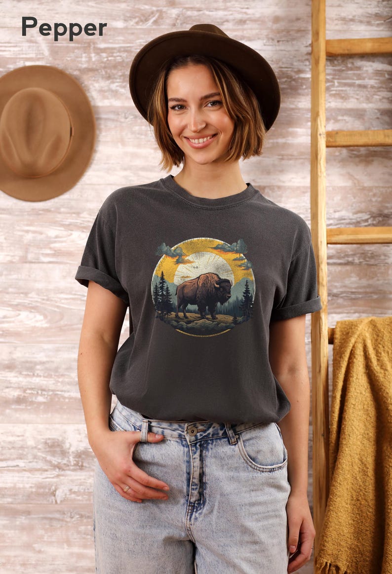 May include: A dark gray t-shirt with a vintage-style graphic of a buffalo standing in a forest with a sunset in the background. The graphic is in a circle with the words "Wild & Free" written below it.