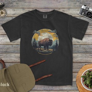 May include: A black t-shirt with a vintage-style graphic of a bison standing in a forest with a sunset in the background.